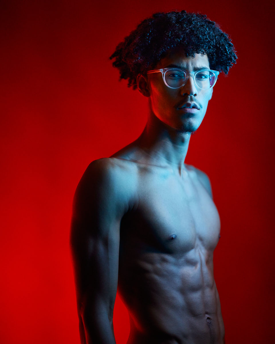 Kasan stands shirtless against a red background with dramatic blue lighting on his body. He is wearing clear glasses, showcasing his muscular physique in a bold and artistic style.