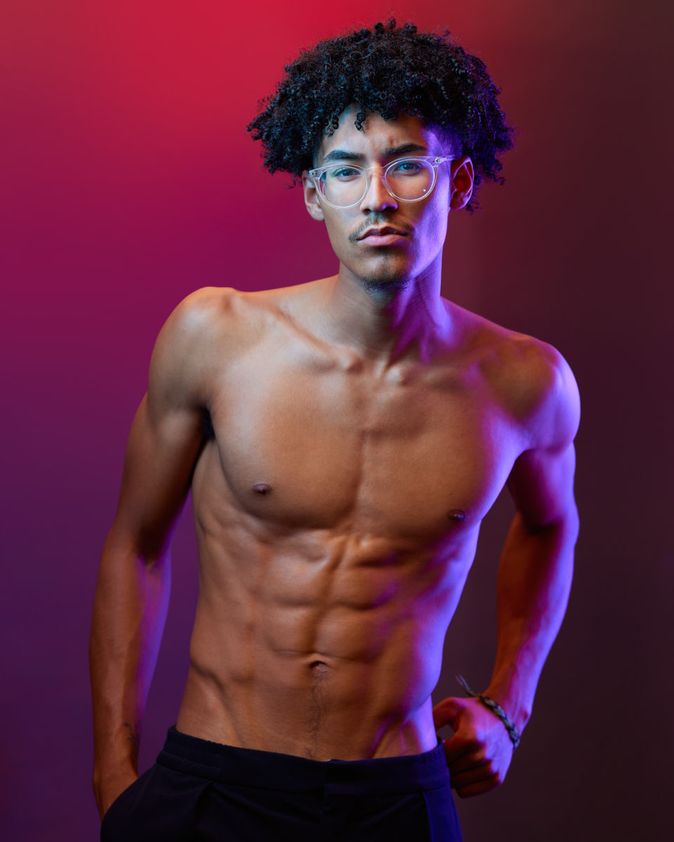 Kasan is shirtless, standing confidently with his hands on his hips against a background with red and purple lighting. His expression is strong and assertive, highlighting his fitness and confidence.