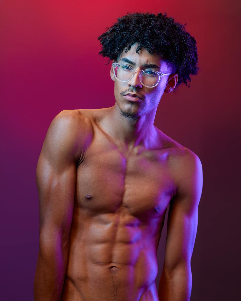 Male modeling portfolio shoot in Chicago-Another shot of Kasan shirtless, wearing clear glasses, standing against a red and purple gradient background. The lighting emphasizes his toned physique and serious expression.