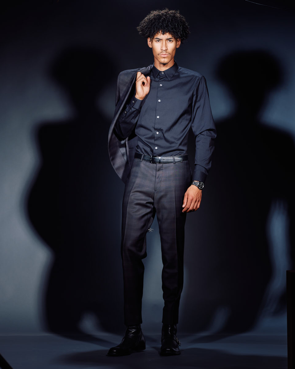 Male modeling portfolio shoot in Chicago-Kasan is wearing a dark suit, holding the jacket over his shoulder, standing against a blue background. The lighting creates a shadow behind him, giving a stylish and confident look.