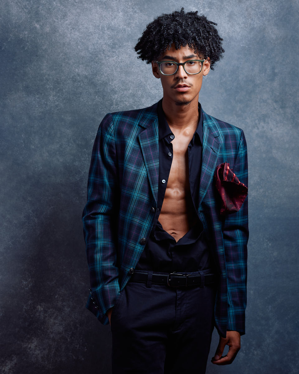 Kasan is wearing a plaid jacket with no shirt underneath, paired with dark trousers. He is posing against a textured grey background, giving a stylish and edgy appearance.