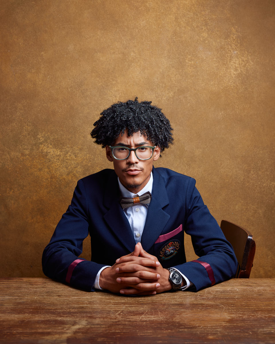 Kasan is seated at a wooden table, wearing a blue blazer with a bow tie and glasses. The background is a warm brown, creating a classic and sophisticated atmosphere.