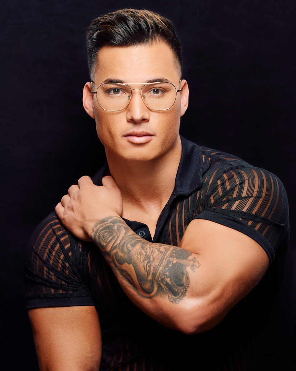 This portrait shows Zah wearing the same striped shirt and glasses, but here, he is resting his arm across his chest, revealing a detailed tattoo on his forearm. The close-up shot highlights both his facial features and the intricate tattoo design.