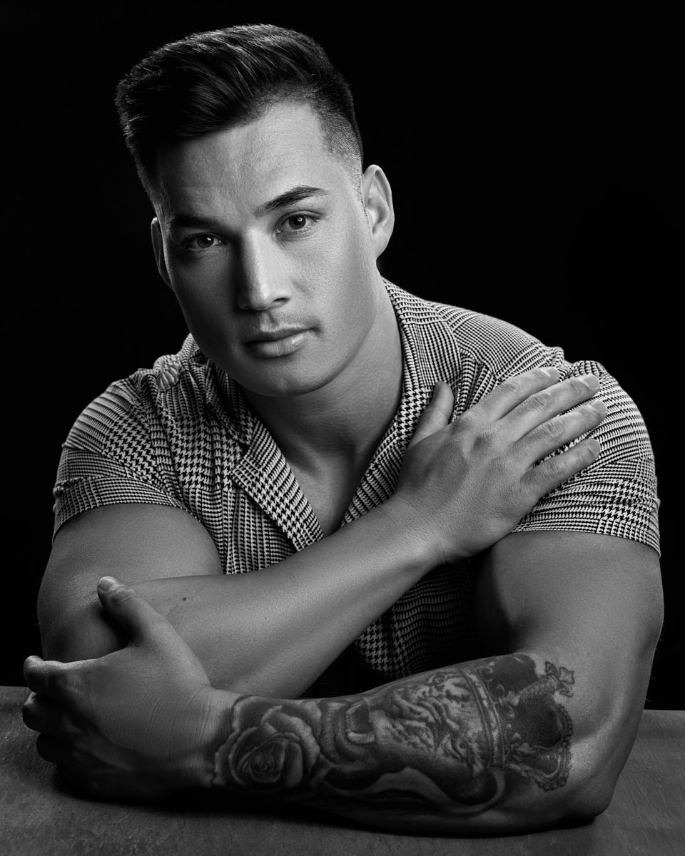 Zah is captured in a black and white portrait, where he is wearing a patterned shirt. His expression is intense, with his arms crossed, showcasing his tattooed arm prominently. The lighting creates deep shadows, emphasizing the contours of his face and the texture of the tattoo.