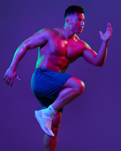 In this dynamic shot, Zah is posing as if in mid-run, wearing blue athletic shorts. The image uses neon or colored lighting, with shades of pink and blue creating a vibrant, energetic effect. The pose, lighting, and colors give the image a modern and artistic look, highlighting Zah’s athletic build and motion.
