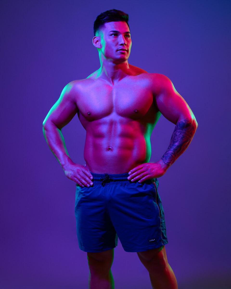 This image shows Zah shirtless in blue athletic shorts, confidently posing with his hands on his hips. The vibrant pink and green lighting highlights his muscular physique against a purple background, emphasizing his strength and focus, while his tattoo adds a personal touch.