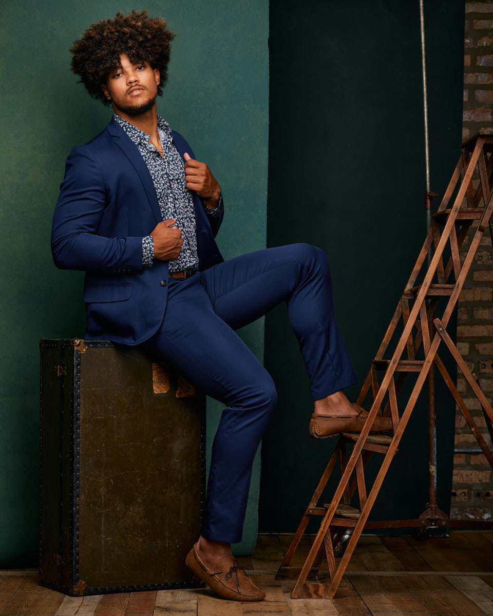 Male Model Comp Card in Chicago-A full-body shot of Josiah in a blue suit with a patterned shirt. He is sitting on a large trunk, with one leg on a wooden ladder, striking a confident pose against a green background.