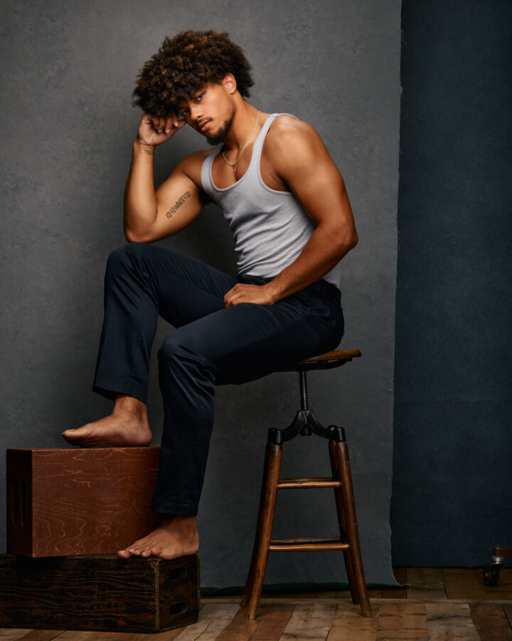 Josiah in a white tank top and dark pants, sitting on a stool with one hand resting on his head and the other on his knee, against a gray background.