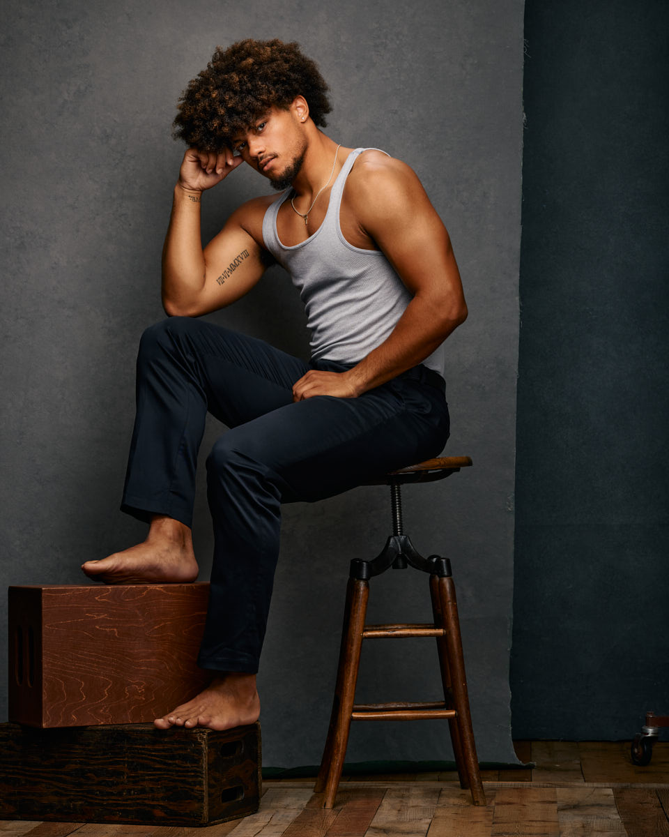 Josiah in a white tank top and dark pants, sitting on a stool with one hand resting on his head and the other on his knee, against a gray background.