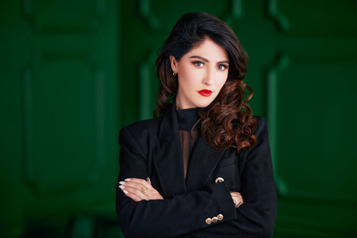 A formal headshot of Catherine Lanigan. She has dark, wavy hair and is wearing a black blazer with gold details. Her red lipstick stands out, and her expression is serious yet confident. The bold green background provides a vibrant contrast to her outfit and appearance, giving her a striking and professional look.