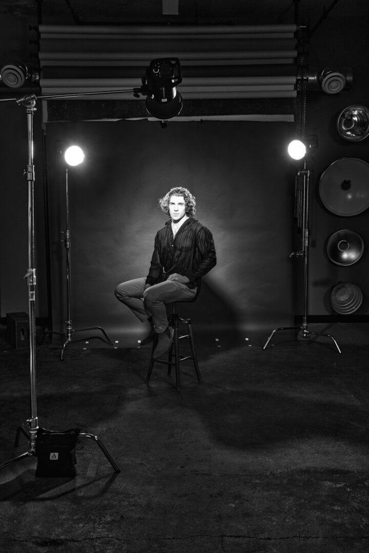A behind-the-scenes studio shot in black and white, showing Xander sitting on a stool under multiple studio lights. The image gives insight into the photoshoot process, with a minimalist backdrop and a candid feel. The lighting is simple, yet it highlights his presence and pose, giving the image a clean, professional look.