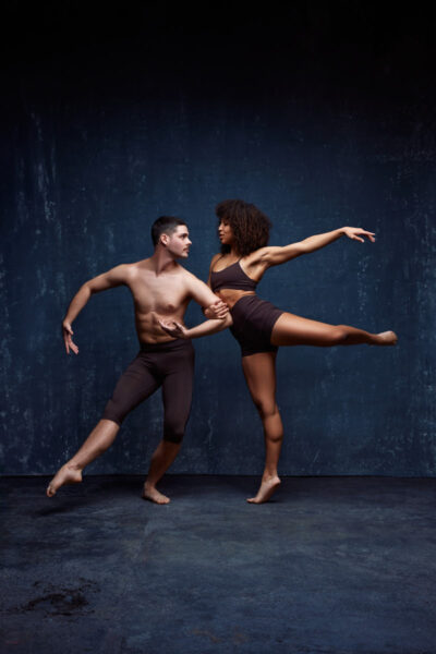 Zach and Mya are in a dynamic dance pose. Zach stands firm, with his arm reaching out toward Mya, who is balanced on one leg, extending the other leg and her arm gracefully. Mya’s pose highlights her flexibility and balance, while Zach supports her with a focused, strong posture. Both wear dark, minimal dance attire, and the lighting emphasizes the clean lines of their bodies against a textured dark backdrop.