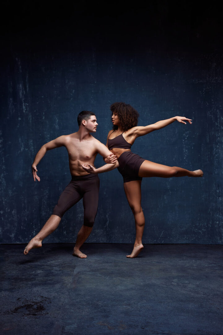 Zach and Mya are in a dynamic dance pose. Zach stands firm, with his arm reaching out toward Mya, who is balanced on one leg, extending the other leg and her arm gracefully. Mya’s pose highlights her flexibility and balance, while Zach supports her with a focused, strong posture. Both wear dark, minimal dance attire, and the lighting emphasizes the clean lines of their bodies against a textured dark backdrop.