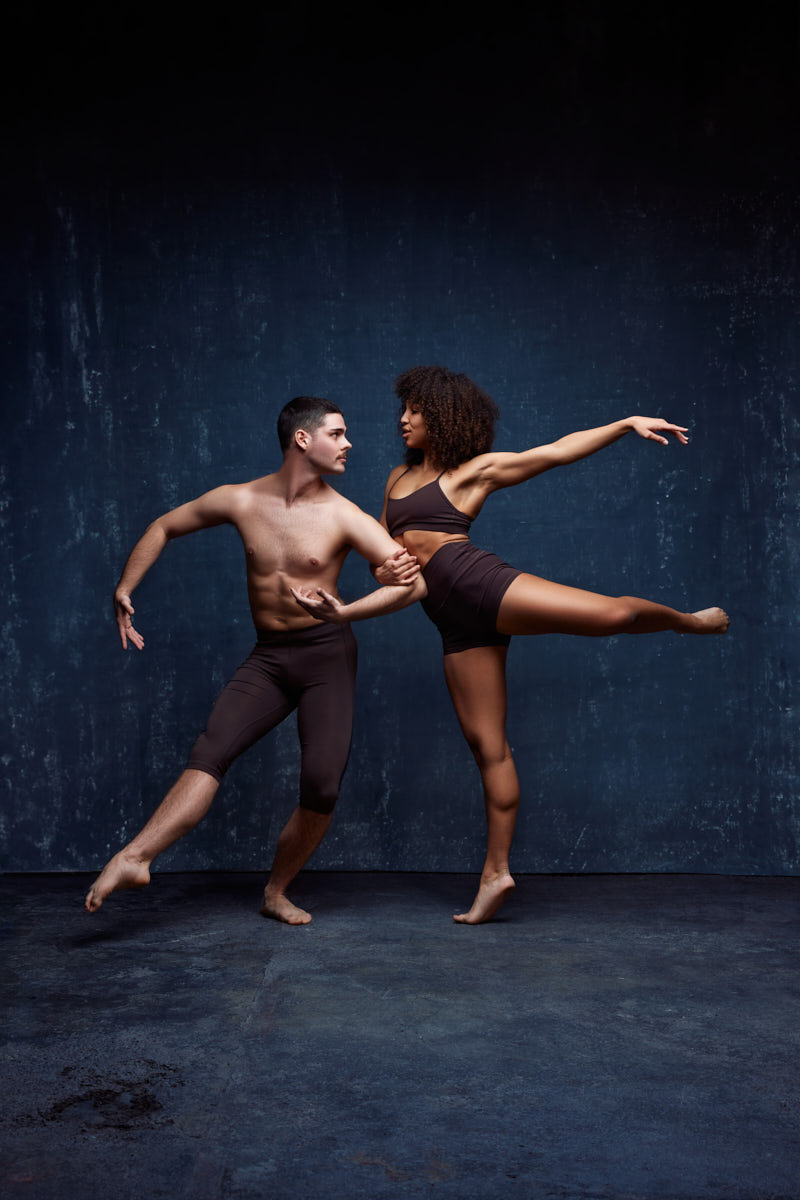 Zach and Mya are in a dynamic dance pose. Zach stands firm, with his arm reaching out toward Mya, who is balanced on one leg, extending the other leg and her arm gracefully. Mya’s pose highlights her flexibility and balance, while Zach supports her with a focused, strong posture.