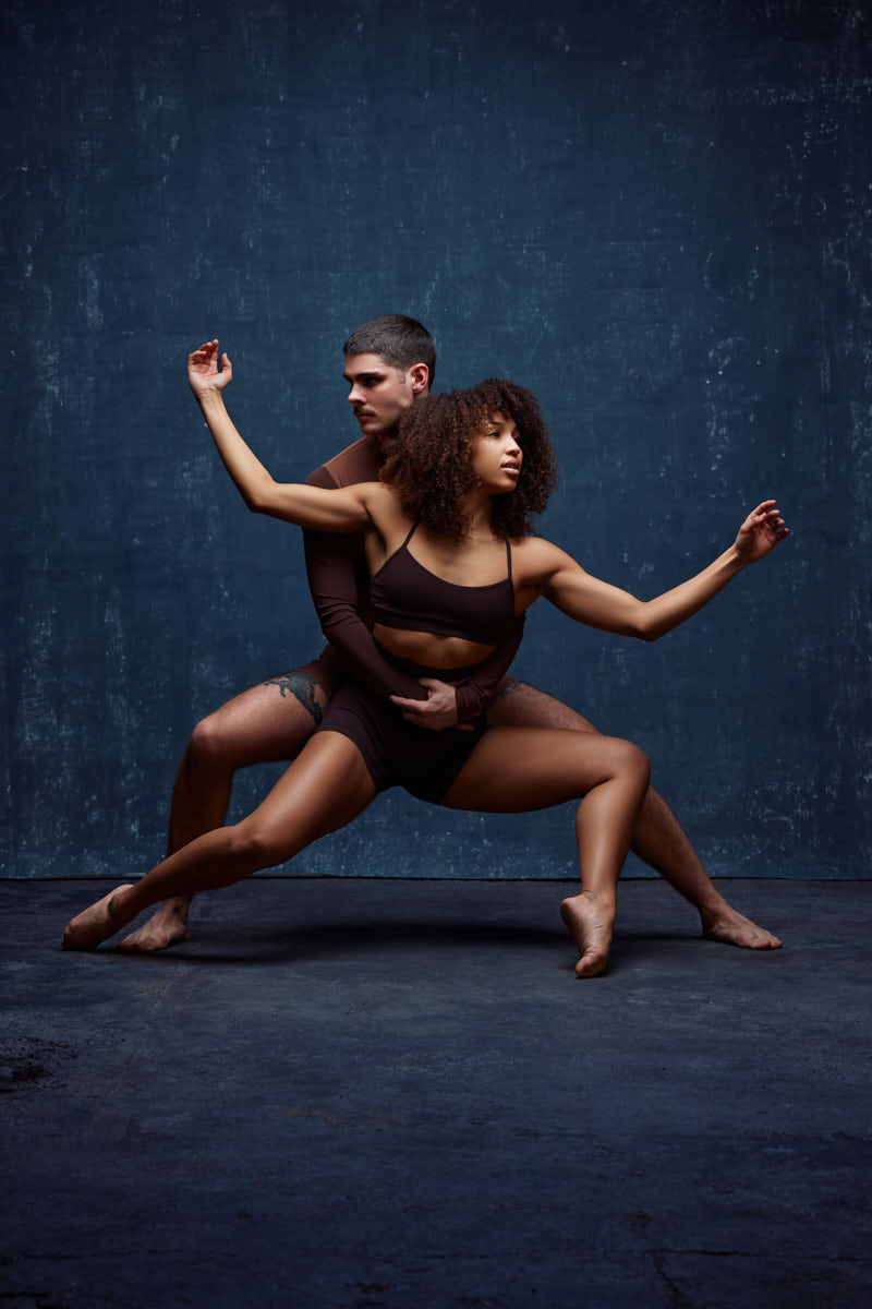 Modern Dance photographer Chicago-Zach and Mya are in a deep, low dance pose, emphasizing both strength and fluidity. Zach is positioned behind Mya, supporting her with his hands as she extends her body in a wide stance.