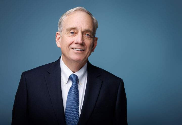 This headshot features a mature gentleman in a formal navy suit, white shirt, and blue tie against a soft blue background. His gentle smile and slightly turned head create a welcoming, professional feel.