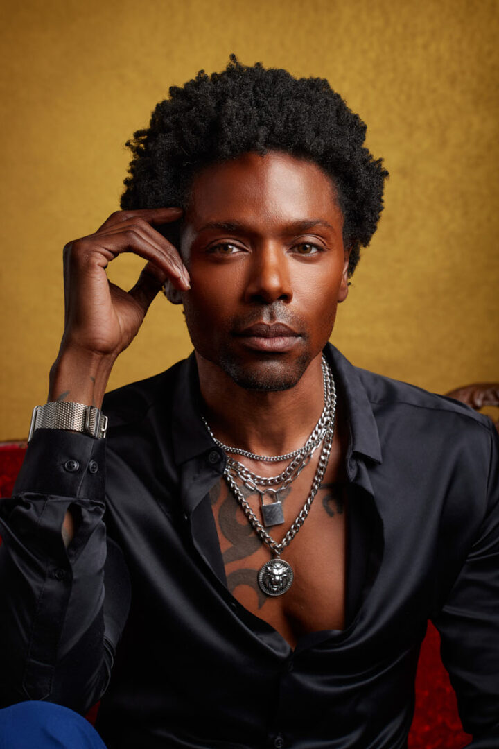 A close-up portrait of Chris on the red couch. His black shirt, layered necklaces, and confident gaze are all in focus, creating a striking portrait against the warm backdrop. His serious expression and carefully styled look emphasize a fashionable, high-end feel.