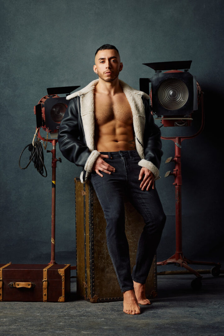 In this cinematic setup, Gael poses shirtless in front of a pair of vintage studio lights. He’s seated on a distressed travel trunk, dressed in fitted black jeans and a two-tone shearling bomber jacket. The lighting and props evoke a nostalgic film set aesthetic, blending fashion and storytelling.