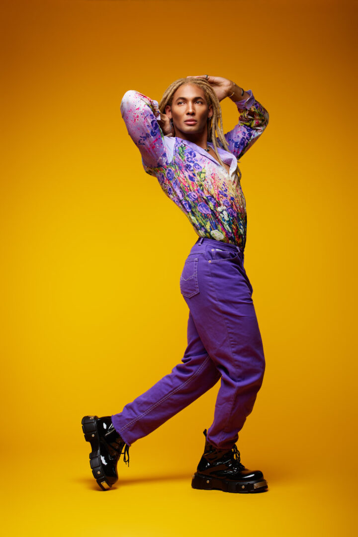Set against a rich yellow background, Chris wears a painterly floral shirt and purple jeans. His curved pose adds dynamic flair, blending movement with elegance—ideal for creative fashion or lifestyle editorials.