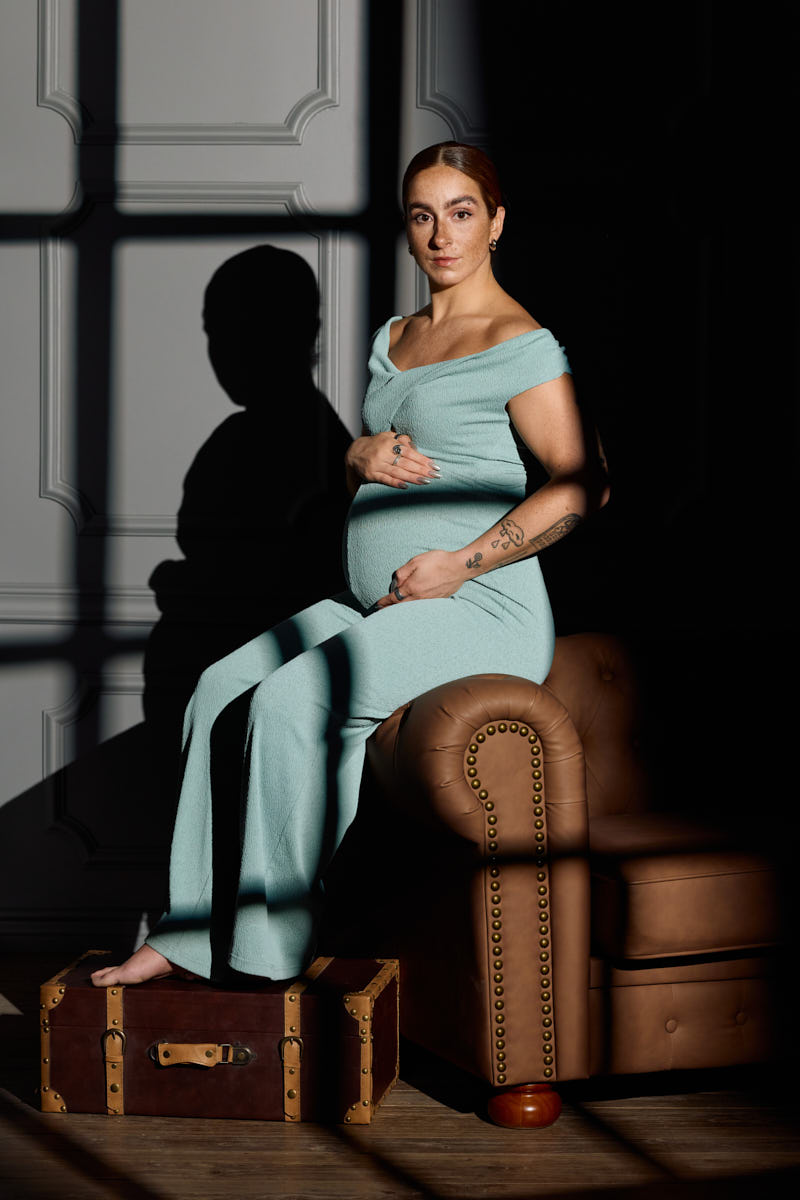 Maternity portrait of a woman seated sideways on a stool, gazing softly at her belly.