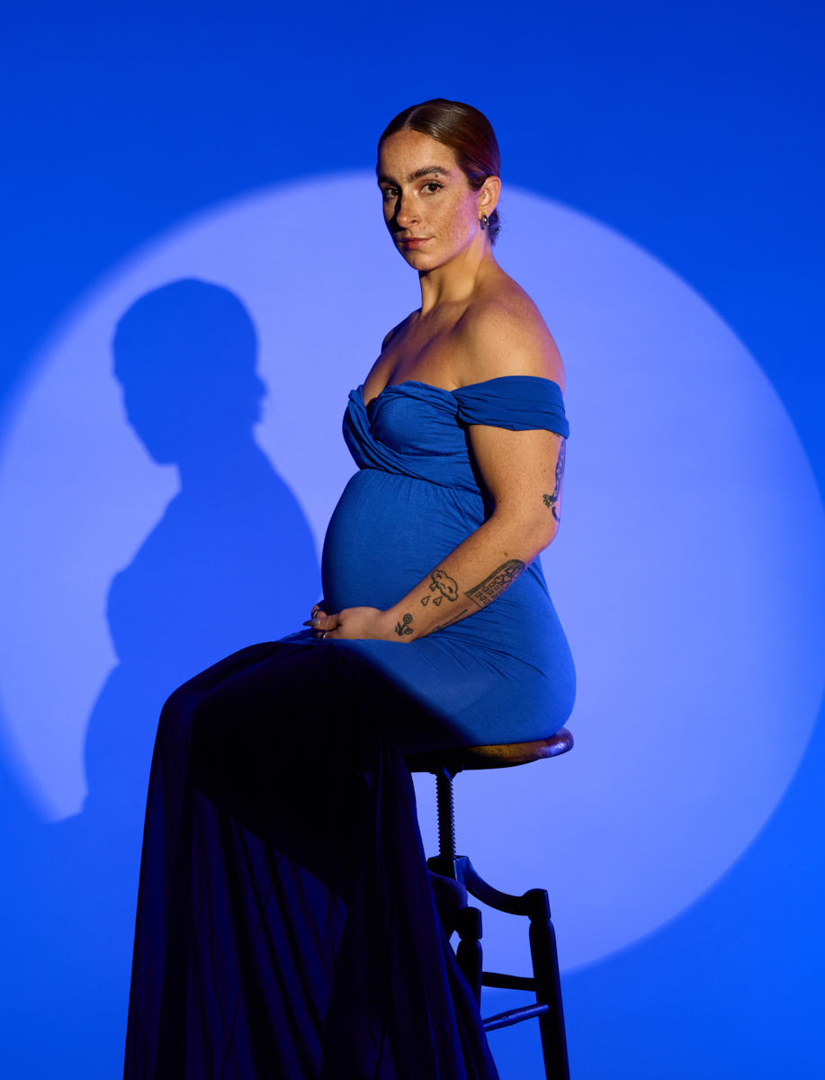 Maternity photo of a woman in a black bodysuit seated on a tall stool, leg raised, and arms at rest.