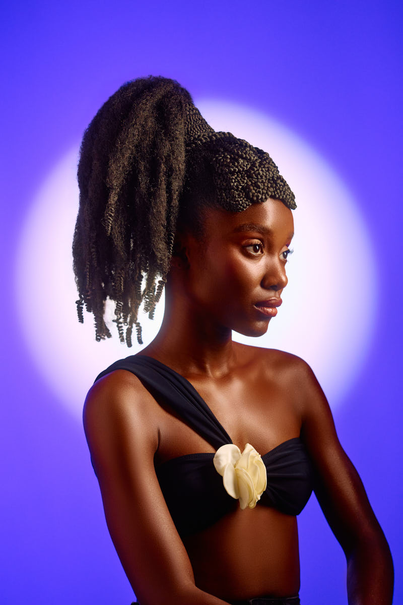 A beautifully composed profile portrait. The model’s long, textured ponytail cascades from an intricate braided crown. She wears a black top with a single strap adorned with a delicate cream flower. The soft halo of light behind her head against the blue background creates an ethereal, graceful feel.