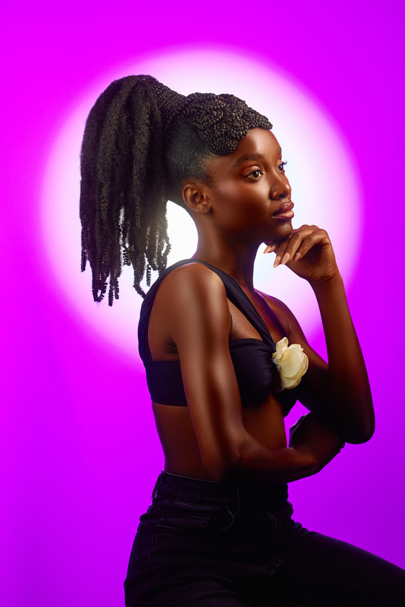 Magazine Editorial Photographer in Chicago-A thoughtful, side-facing portrait of the model seated, resting her chin on her hand in a classic, contemplative pose. Her ponytail and braided crown remain the focal point, complemented by the subtle flower detail on her top. The glowing pink and purple background adds warmth and dreaminess to this editorial-style shot.