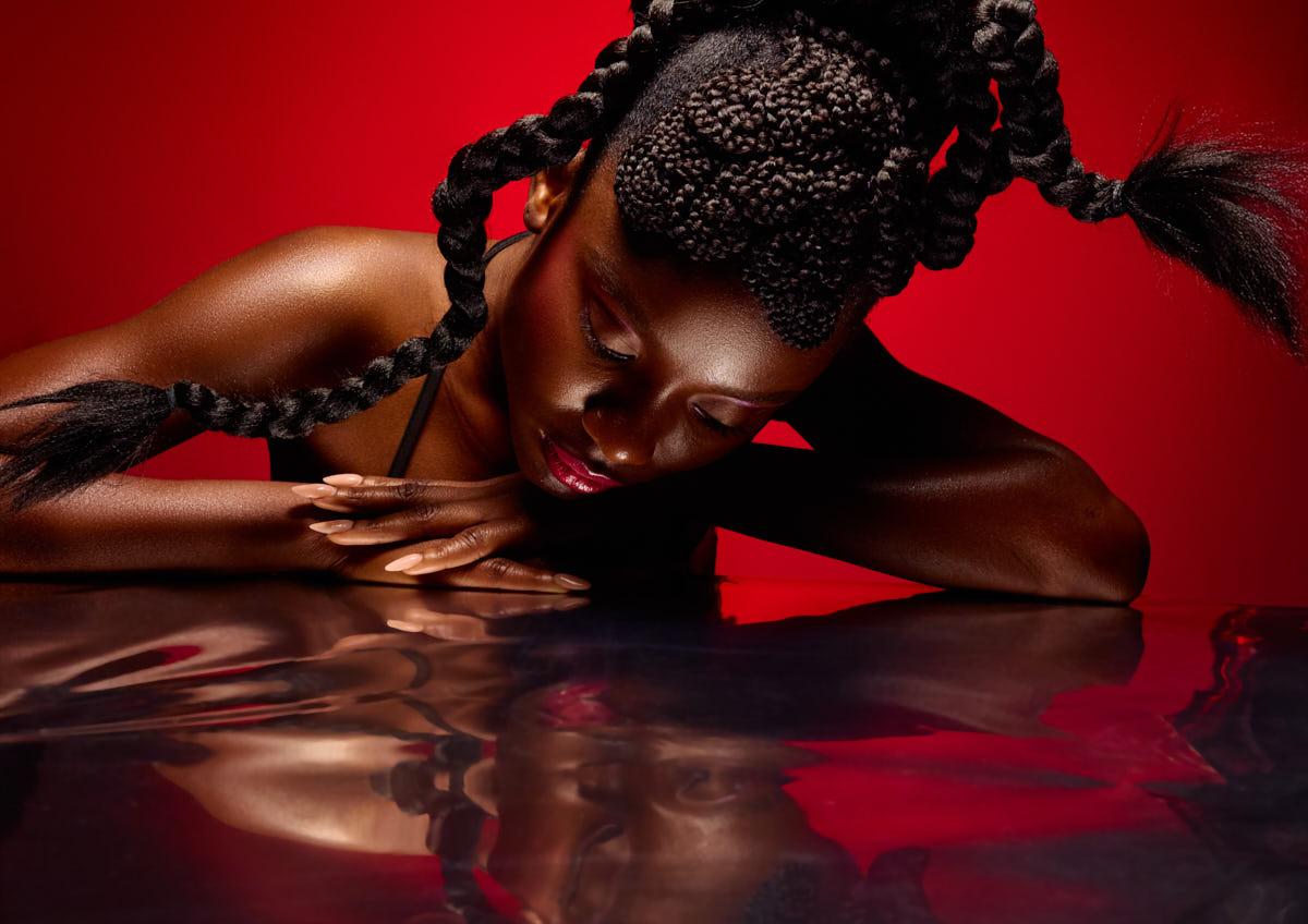 Magazine Editorial Photographer in Chicago-A striking close-up of a model resting her head and arms on a reflective surface. Her hair is intricately braided and styled into bold shapes that fan out dramatically. The vivid red background intensifies the powerful, almost introspective mood. Her reflection in the surface adds depth and an artistic edge.