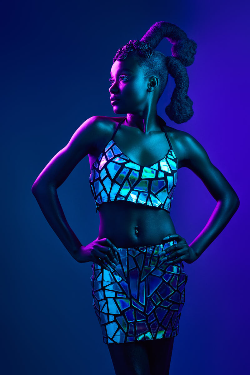 A bold, full-body portrait of the model in a two-piece mirrored outfit that reflects blue and purple hues. She’s standing with her hands on her hips, gazing to the side with confidence. The deep blue background gives this shot a futuristic, editorial look that feels ready for a magazine spread.