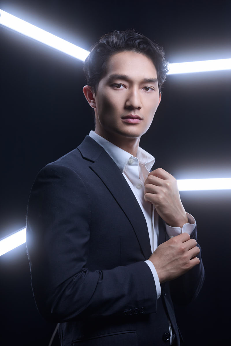 A polished head-and-shoulders shot. The model wears a suit and white shirt, gently adjusting his cufflinks. He stands against a dark background with crisp, futuristic white light beams behind him, creating an upscale, modern energy.