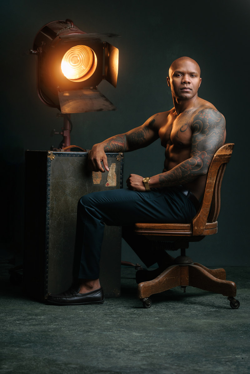 Lee Smith sits shirtless in a vintage wooden chair beside a large studio spotlight. He wears tailored black trousers and loafers, with a gold watch adding a refined touch. His muscular physique and full tattoo sleeve are prominently displayed, and his calm, confident expression creates a powerful editorial presence.