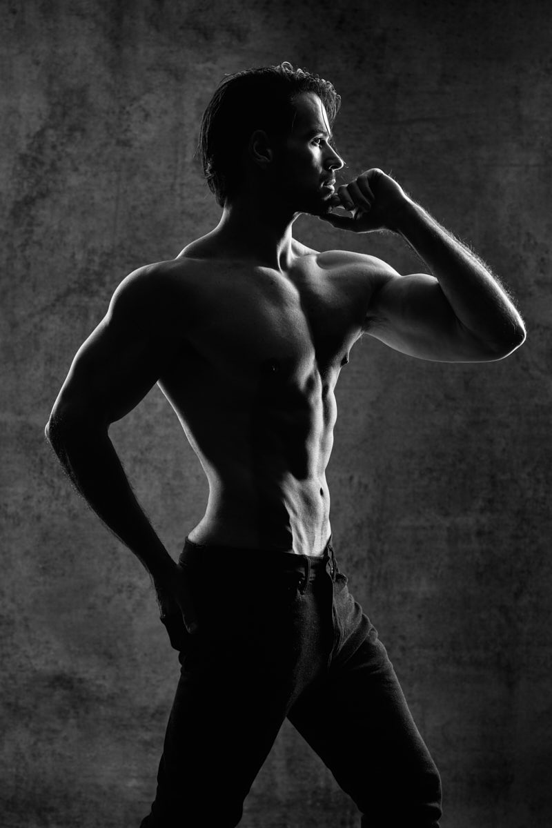 A black-and-white portrait of a shirtless male model standing in profile. One hand rests thoughtfully near his chin while the other sits on his hip. His muscular definition is sharply outlined, and the mood is serious and introspective, giving the image a timeless, classic look.