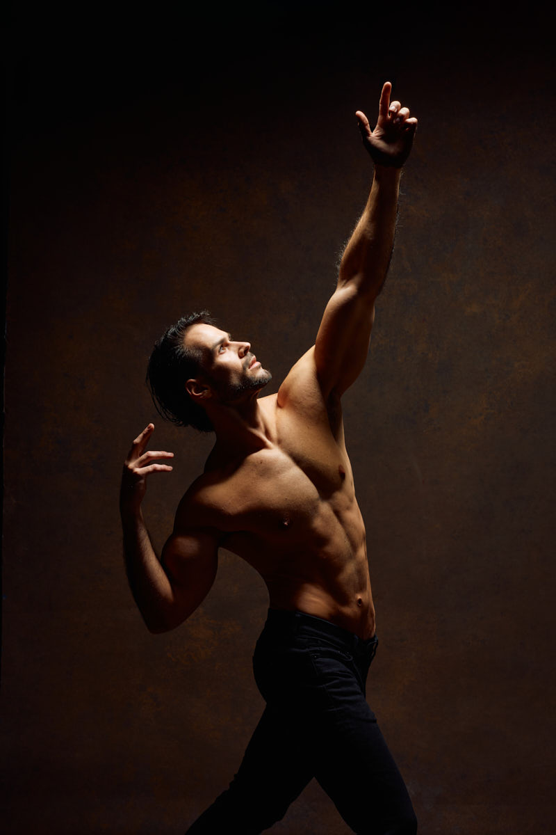 A shirtless male model poses against a warm brown textured backdrop. His body is angled in profile while one arm reaches upward and the other curves gracefully behind him. The pose feels expressive and almost dance-inspired, highlighting his toned torso and athletic physique.
