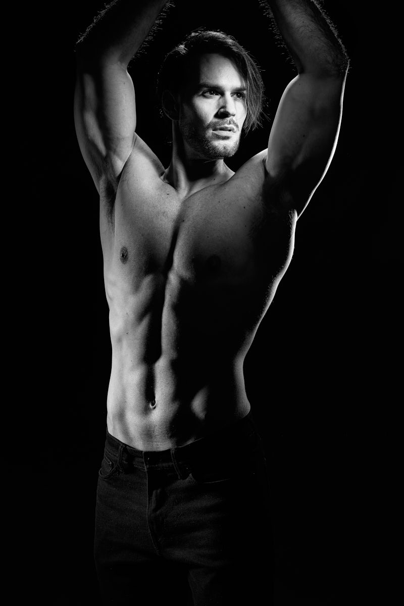 Another black-and-white portrait of the same model, now facing slightly toward the camera with both arms raised overhead. His torso and abdominal definition are prominently featured. His expression is intense and focused, and the overall feel is bold, high-fashion, and powerful.