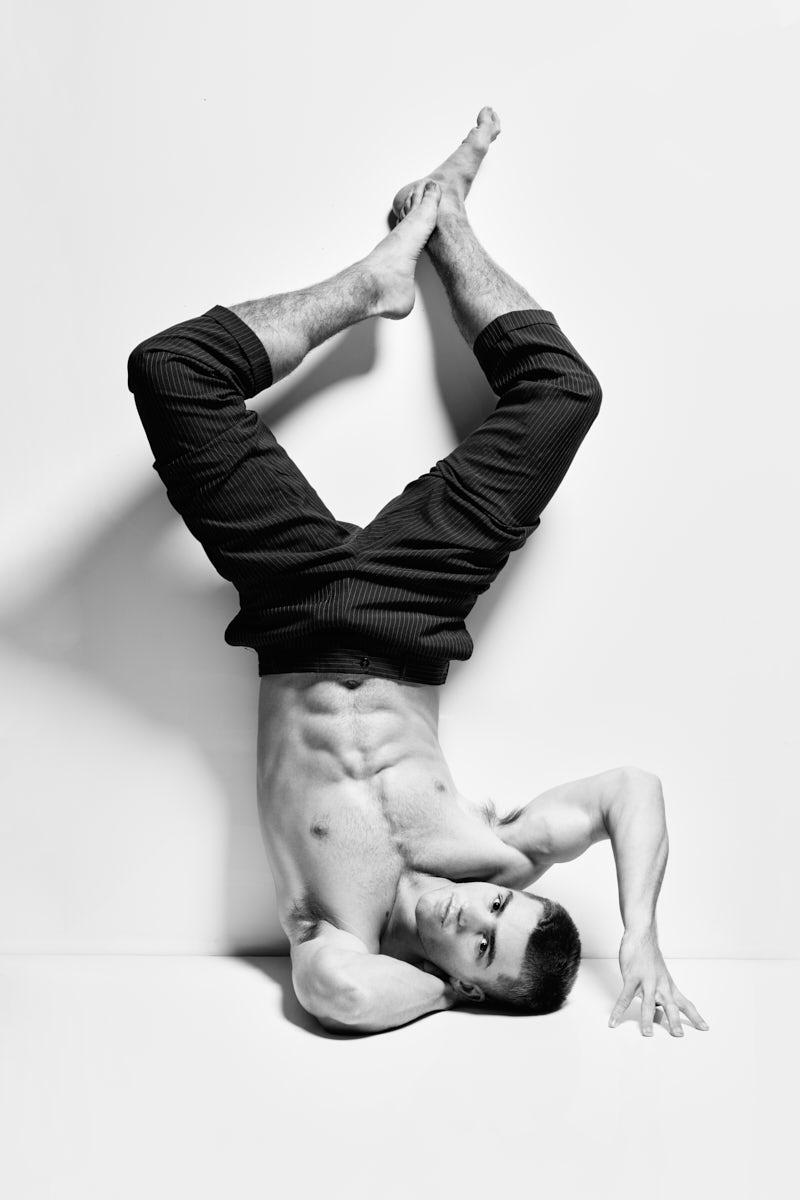 A black-and-white image of a male model posing upside down against a wall. His legs are extended upward while his torso twists slightly, creating a striking, unconventional composition. The photo feels artistic and editorial.