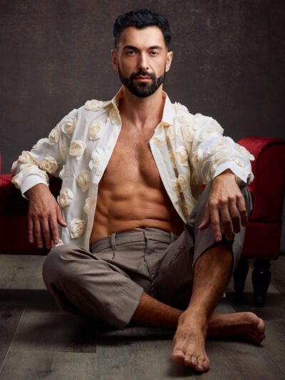 A male model sits on the floor leaning back against a red couch. He wears an open cream-colored shirt decorated with floral details, paired with tailored trousers. His shirt is partially unbuttoned, revealing his torso. His expression is direct and confident, blending sensuality with high-fashion styling.