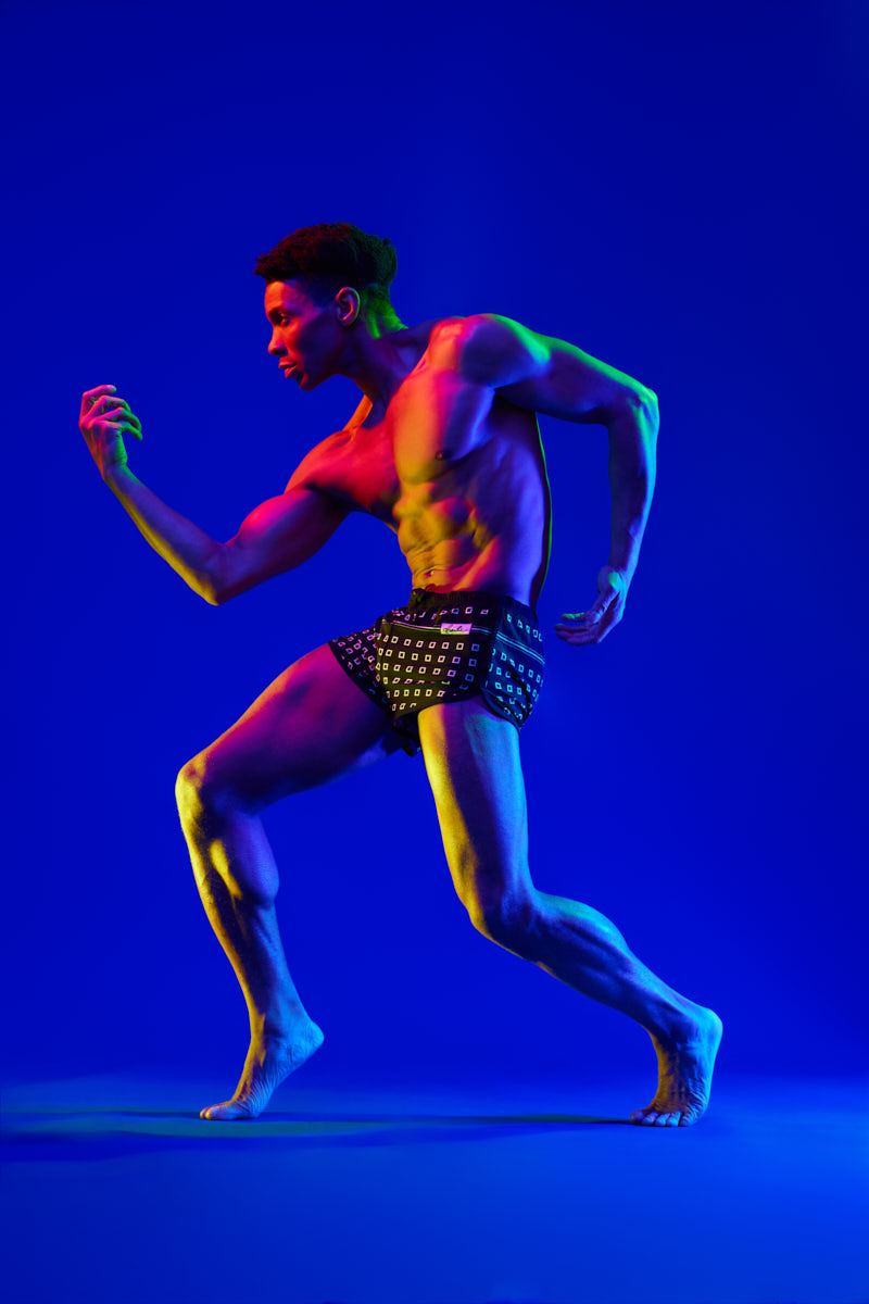 A male model is captured mid-motion against a vivid blue background. Colored light accents his muscular physique as he leans forward in a dynamic pose, wearing fitted shorts. His body position suggests strength and movement, creating a high-impact fitness portrait.