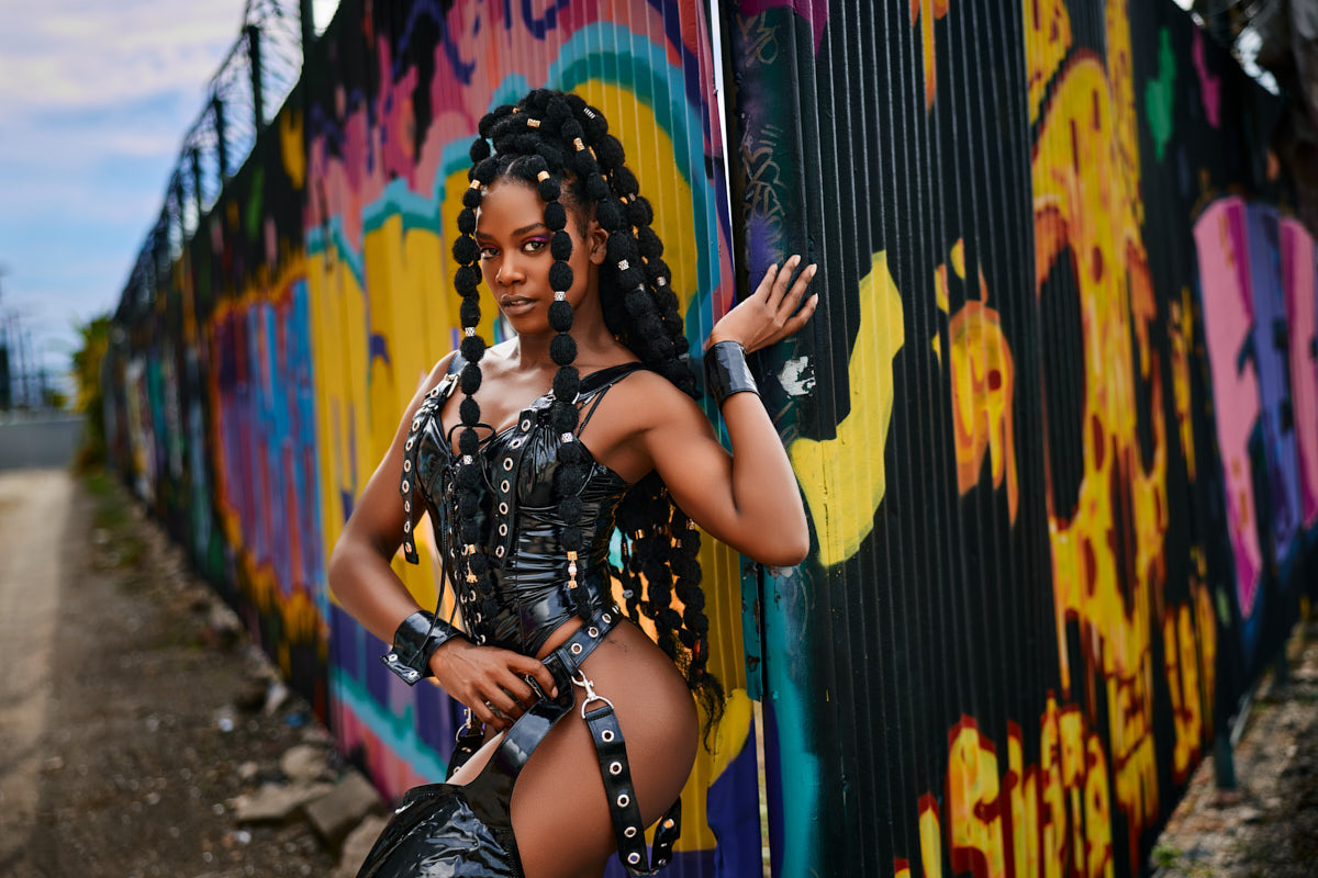 The same female model stands confidently against a colorful graffiti wall, leaning slightly against a metal post. Her braided hairstyle and structured black outfit enhance the edgy, fashion-forward aesthetic.