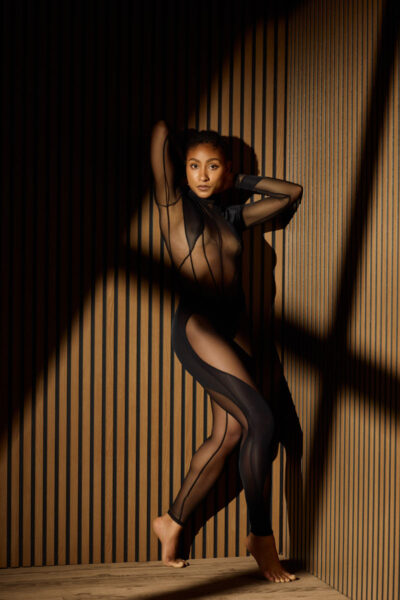 Valeriane stands barefoot against a vertical wooden slat wall, wearing a sheer black bodysuit with sculptural cutouts that contour her silhouette. Her pose is elongated and fluid, one arm raised above her head while the other frames her body, creating elegant lines from shoulder to toe. Dramatic shadows fall across the wall and her figure, emphasizing the architectural quality of the setting. The image feels high-fashion, refined, and powerful, with a strong editorial presence.