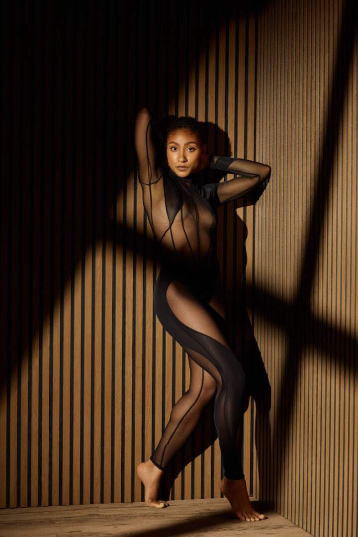 Valeriane stands barefoot against a vertical wooden slat wall, wearing a sheer black bodysuit with sculptural cutouts that contour her silhouette. Her pose is elongated and fluid, one arm raised above her head while the other frames her body, creating elegant lines from shoulder to toe. Dramatic shadows fall across the wall and her figure, emphasizing the architectural quality of the setting. The image feels high-fashion, refined, and powerful, with a strong editorial presence.