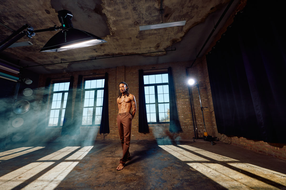 Athletic male trainer-style portrait walking barefoot through an industrial studio with tall windows and long shadows