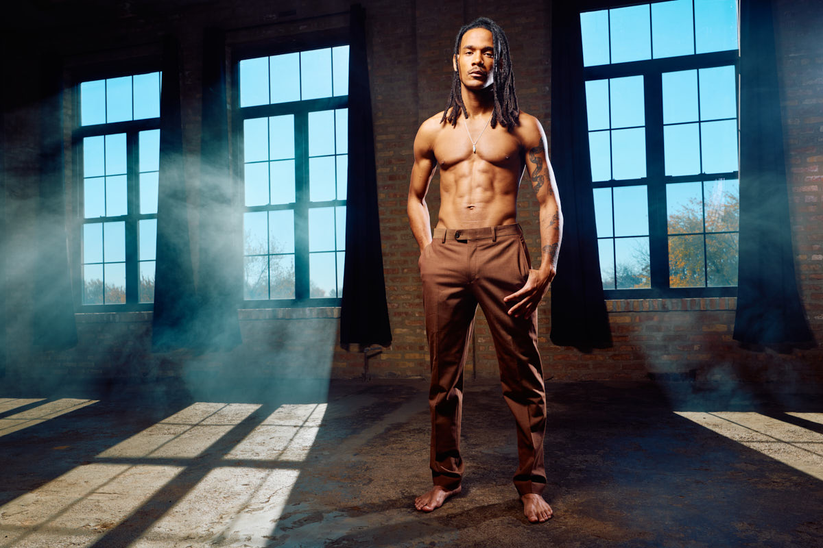 Shirtless athlete standing confidently in an industrial studio with tall windows and dramatic shadows