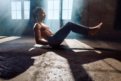 The model is seated on the studio floor, leaning back slightly with his legs lifted and bent at the knees. His abdominal muscles are engaged, showcasing strength and control. The directional light from the windows creates striking shadows, and a faint mist in the air adds dimension to the industrial setting.