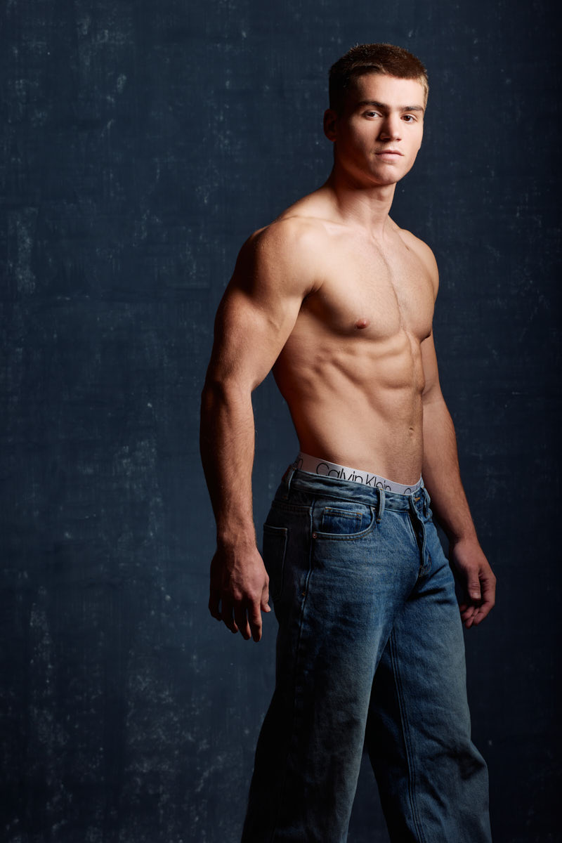 Profile fitness portrait of a shirtless male in jeans against a blue background showing shoulder and arm definition