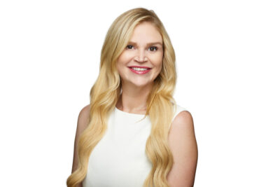A blonde woman in a sleeveless cream top poses against a clean white background. Her long, softly styled hair frames her face, and she smiles warmly at the camera. The neutral background gives the portrait a bright, modern, and professional feel, perfect for business websites or LinkedIn profiles.