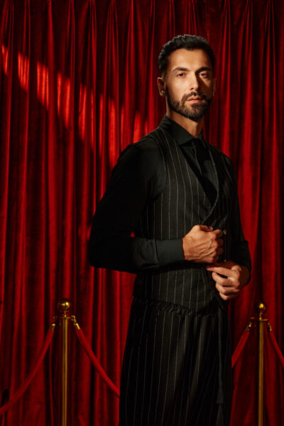 The same male model stands confidently in front of rich red velvet curtains with gold stanchions in the foreground, evoking a dramatic, theatrical atmosphere. He wears a black dress shirt paired with a pinstripe vest, adjusting it slightly in a composed gesture. His posture is upright and poised, projecting authority and sophistication. The lighting emphasizes the luxury of the setting, making this portrait feel bold, cinematic, and reminiscent of a premiere or upscale event setting.