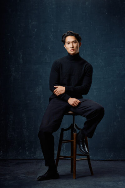 The final image presents a refined studio portrait of the model seated on a wooden stool against a textured blue background. He wears a black turtleneck sweater and tailored trousers, creating a clean and minimalist fashion look. His relaxed posture and calm expression contrast with the dramatic lighting, resulting in a sophisticated and modern fashion portfolio image.
