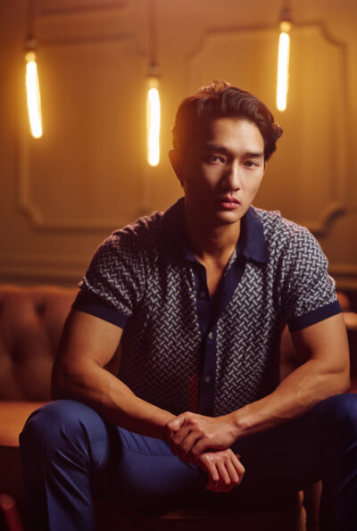 A stylish portrait of a male model seated on a leather couch in a softly lit interior. Warm pendant lights glow behind him, adding depth and atmosphere. He wears a patterned short-sleeve shirt and dark trousers, leaning forward slightly with an intense, serious expression. The overall mood feels cinematic and upscale.