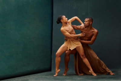 The dancers reconnect in a sculptural partnered pose where the female dancer lifts her torso and arm upward while the male dancer anchors her movement with a firm grip. Their expressions convey concentration and intensity, capturing the emotional storytelling often found in contemporary dance performance.