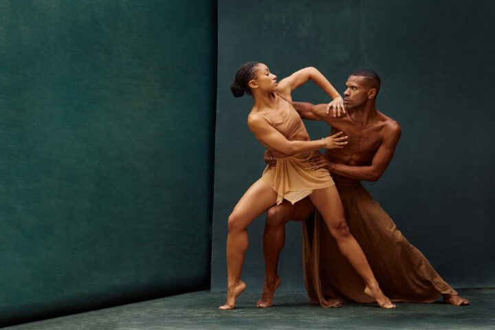The dancers reconnect in a sculptural partnered pose where the female dancer lifts her torso and arm upward while the male dancer anchors her movement with a firm grip. Their expressions convey concentration and intensity, capturing the emotional storytelling often found in contemporary dance performance.